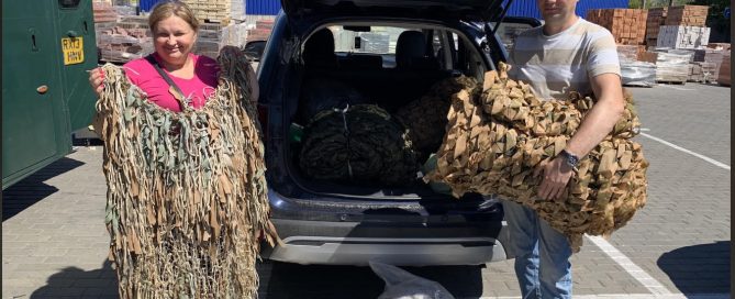 Two volunteers holding camouflage netting beside an SUV with the trunk open in a parking lot