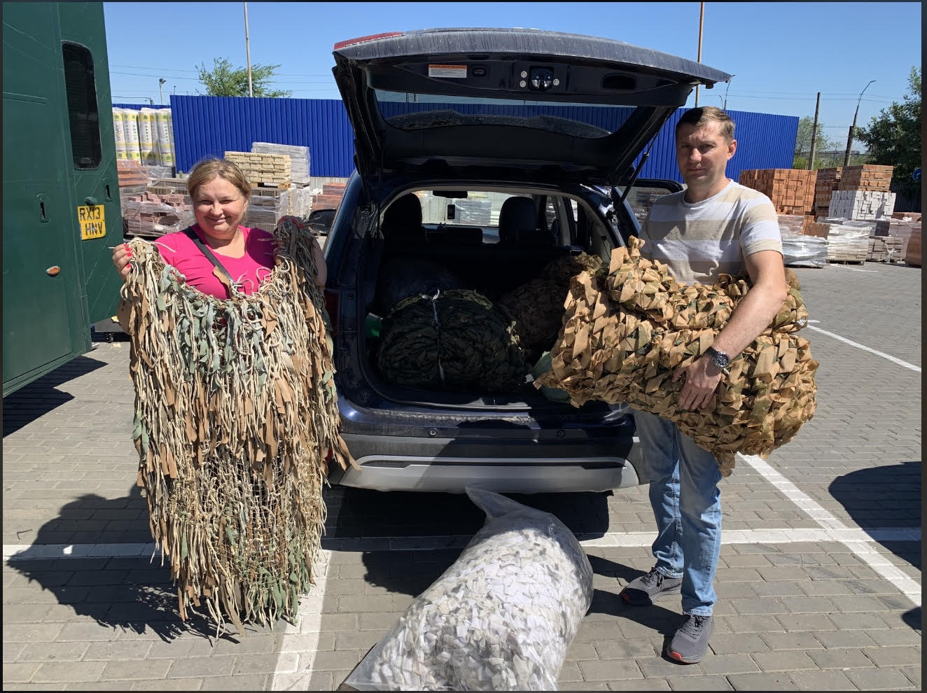 Two volunteers holding camouflage netting beside an SUV with the trunk open in a parking lot